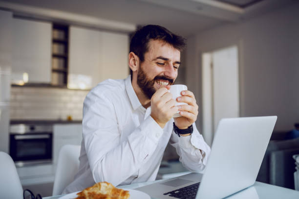 Young cheerful smiling attractive bearded man sitting at dinning table, reading an e-mail on laptop and enjoying his fresh morning coffee.